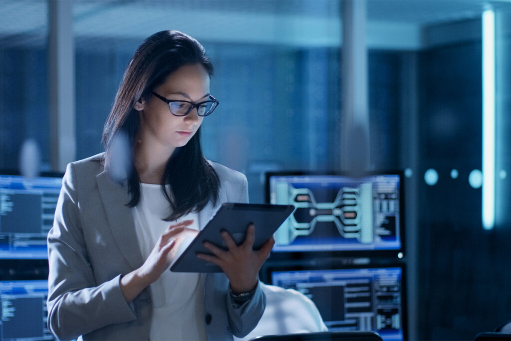 Young Female Government Employee Wearing Glasses Uses Tablet in System Control Center. In the Background Her Coworkers are at Their Workspaces with many Displays Showing Valuable Data.